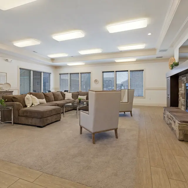 A spacious living room featuring a large sectional sofa, a coffee table, and two armchairs. The room has several windows allowing natural light and has a stone fireplace with a television mounted above it.