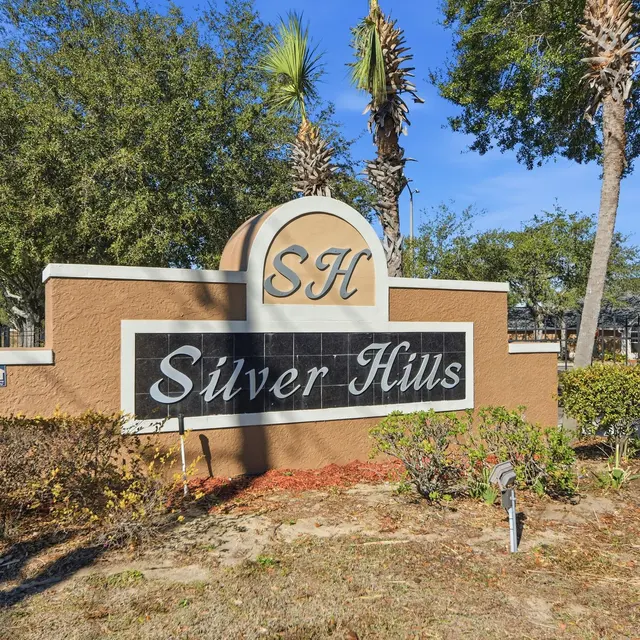 A decorative sign for 'Silver Hills' featuring the initials 'SH' on top, surrounded by greenery and a clear blue sky.
