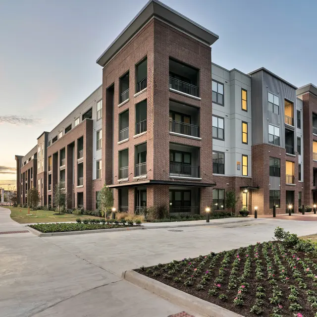Modern Apartment Complex at Dusk A modern apartment complex with a combination of brick and gray exterior. The structure showcases multiple levels, with balconies visible on several units. Landscaping with flowers and well-maintained pathways are visible in the foreground, set against a sunset sky.