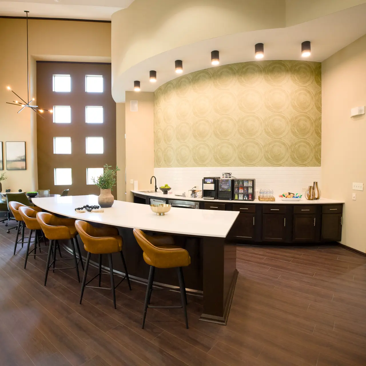 A modern office kitchen area featuring a long countertop with stools, a coffee machine, and cabinetry. Large windows provide natural light in a stylish, ergonomic design.
