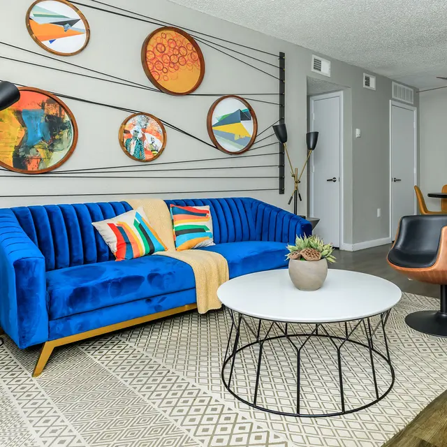 A vibrant modern living room featuring a blue velvet sofa with colorful pillows, a round white coffee table, unique wall art, and stylish dining area.
