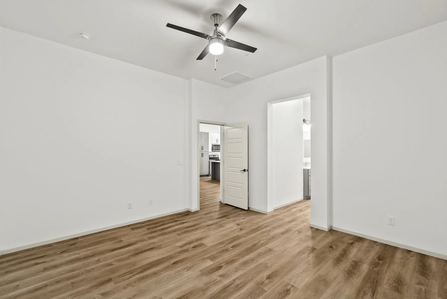 A spacious, modern room with light walls, wooden flooring, a ceiling fan, and an open doorway leading to another area.