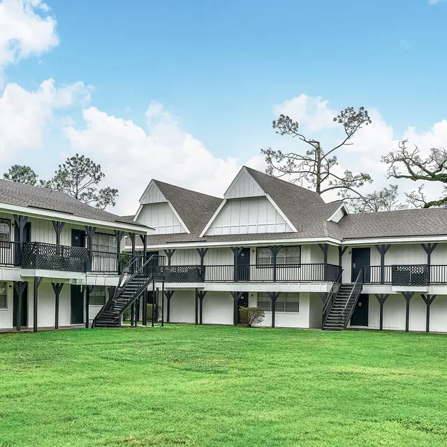 A view of a multi-story apartment complex set in a green grassy area under a bright blue sky with a few clouds. The buildings feature a white exterior with black railings and multiple staircases.