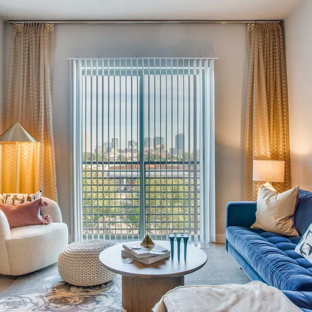 A modern living room with large windows providing a view of the city. It features a blue velvet sofa, a white armchair, a round coffee table, and decorative art on the walls. Soft lighting creates a cozy atmosphere.
