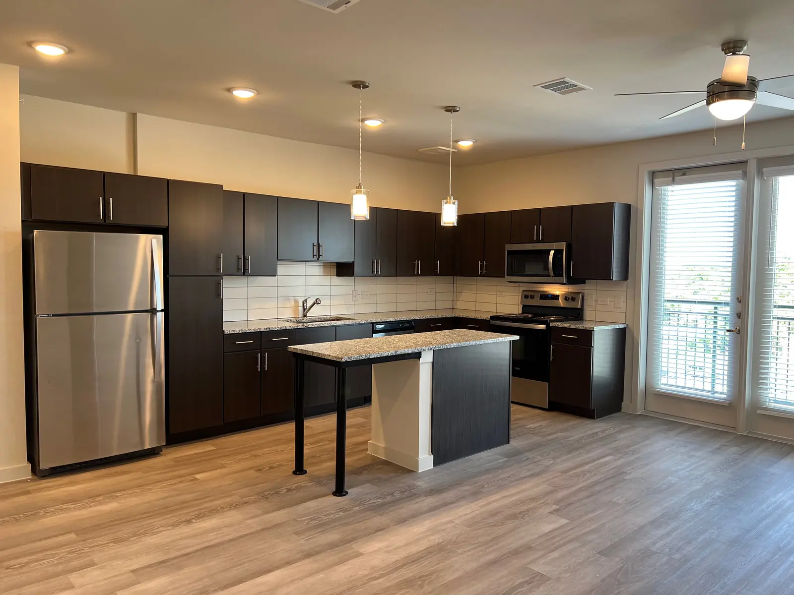 A modern kitchen featuring dark cabinetry, stainless steel appliances, and a granite countertop with a small island. It has large windows that let in natural light, illuminating the open floor plan with light-colored wooden flooring.