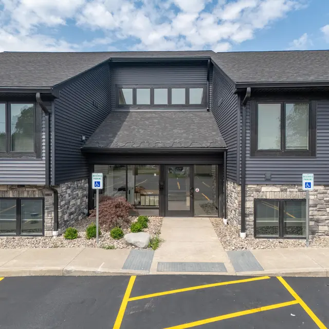A modern building exterior featuring a dark blue and stone facade with a welcoming entrance. The parking area is visible in the foreground with marked spaces. Clear skies overhead add to the bright atmosphere.