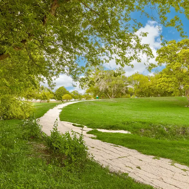 A scenic view of a winding pathway in a park surrounded by trees and lush green grass under a blue sky with clouds.