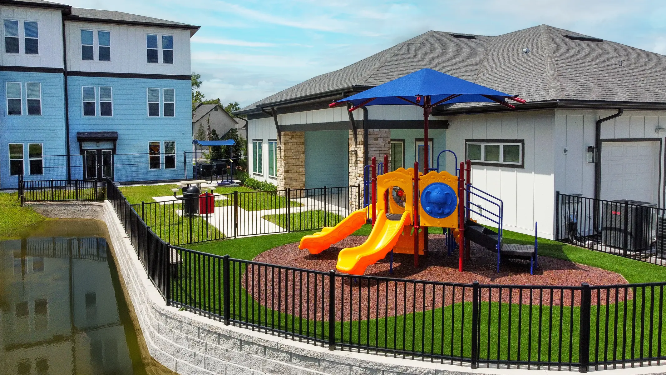 A colorful playground with slides surrounded by a fence, next to a building. The area features green grass and playground equipment under a blue shade.