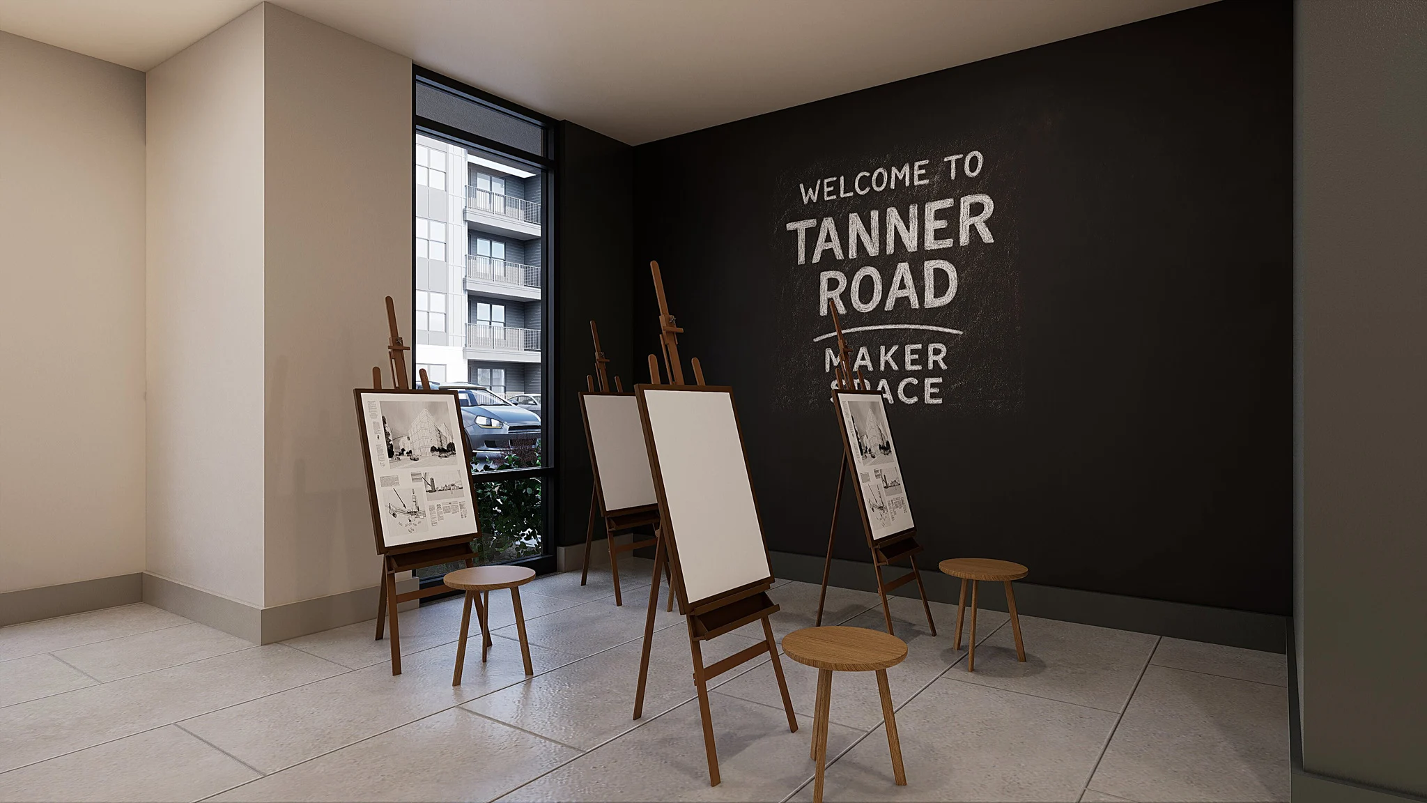 An interior space with easels and stools set up for creative activities. A large chalkboard wall displays 'WELCOME TO TANNER ROAD MAKER SPACE.' Natural light enters from a window, highlighting the modern design.