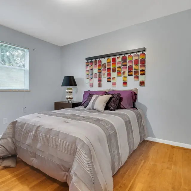 A cozy bedroom featuring a bed with gray bedding and purple pillows, a modern wall hanging above the bed, and a window with blinds.