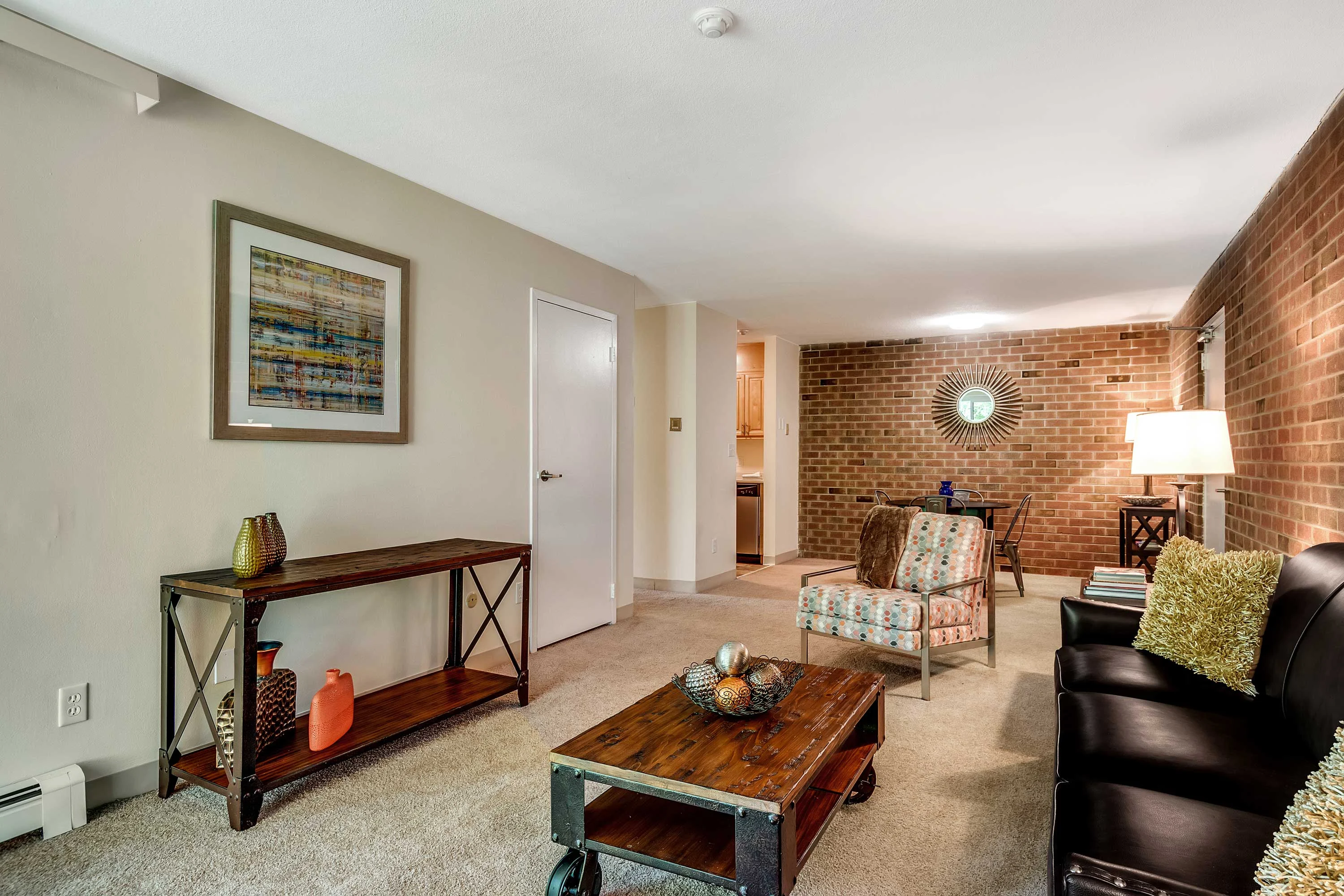 A cozy living room featuring a brown leather sofa, a patterned chair, a wooden coffee table, and a side table with a lamp. The walls are painted beige with a brick accent wall, and there's a colorful artwork hanging on the wall.
