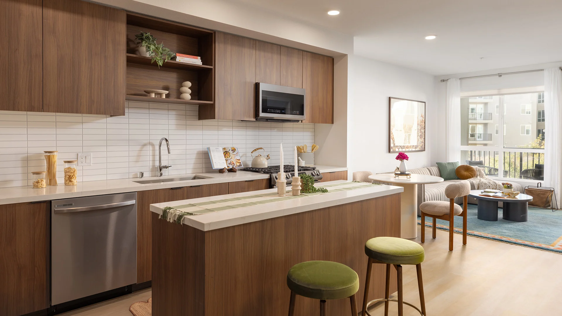 Contemporary Kitchen and Living Room Design A modern kitchen with wooden cabinets, a stainless steel dishwasher, and an island with green stools. The kitchen features a gray countertop and a tiled backsplash, with a comfortable living area visible in the background, showcasing large windows and contemporary furniture.