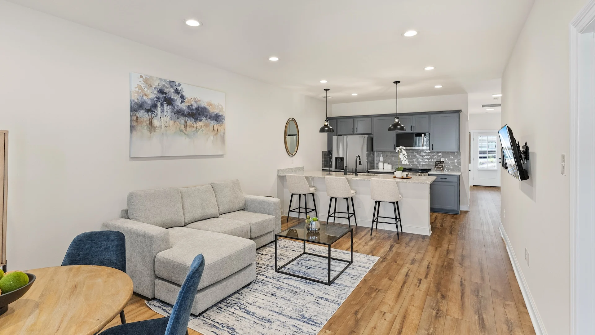 Contemporary Open-Concept Living Space A modern, open-concept living area featuring a light gray sofa, a wooden dining table with blue chairs, and a well-equipped kitchen with bar seating.