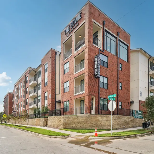 A modern multi-unit apartment building with a brick façade and balconies, located on a sunny street with clear blue skies.
