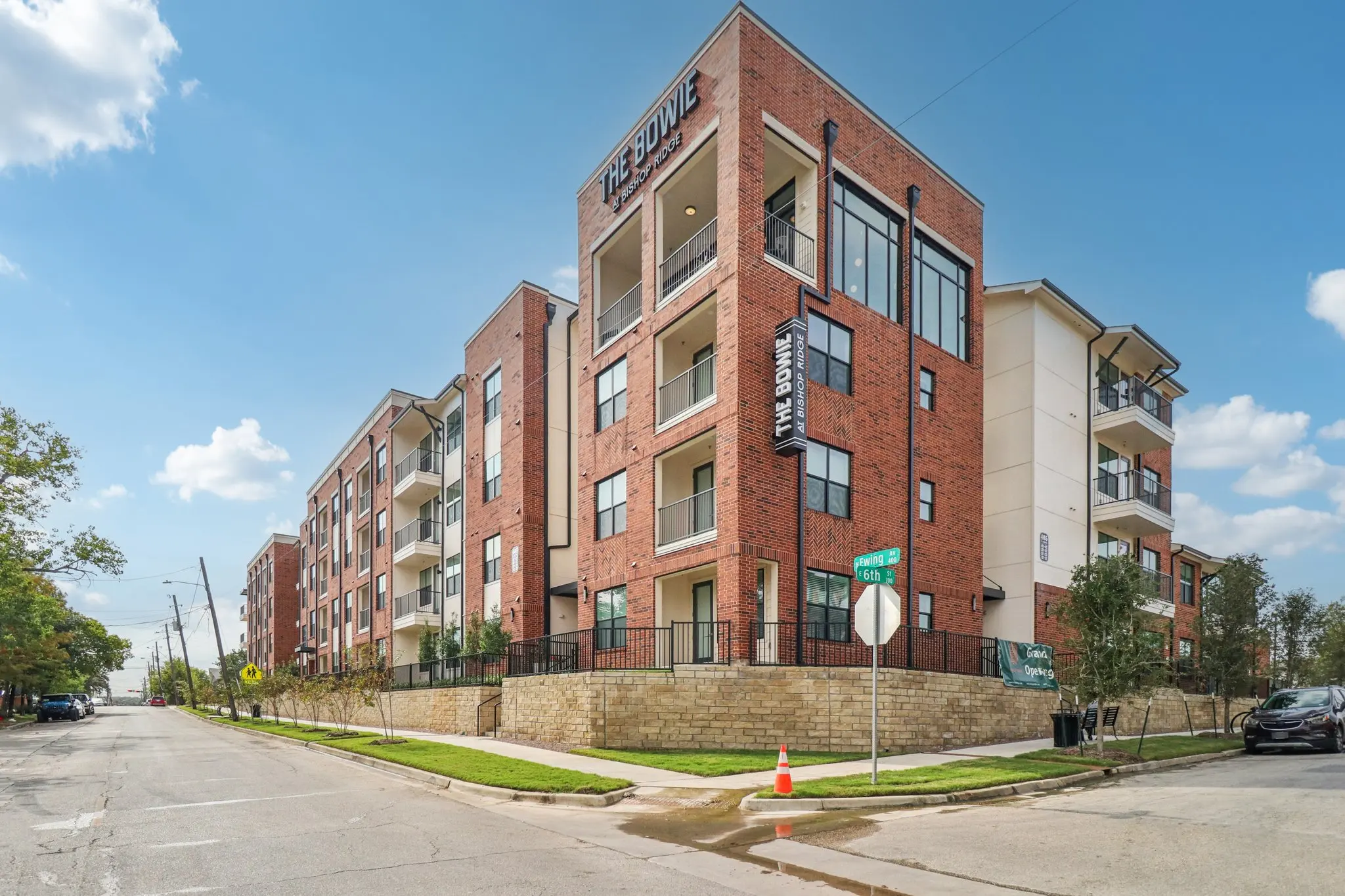 A modern multi-unit apartment building with a brick façade and balconies, located on a sunny street with clear blue skies.