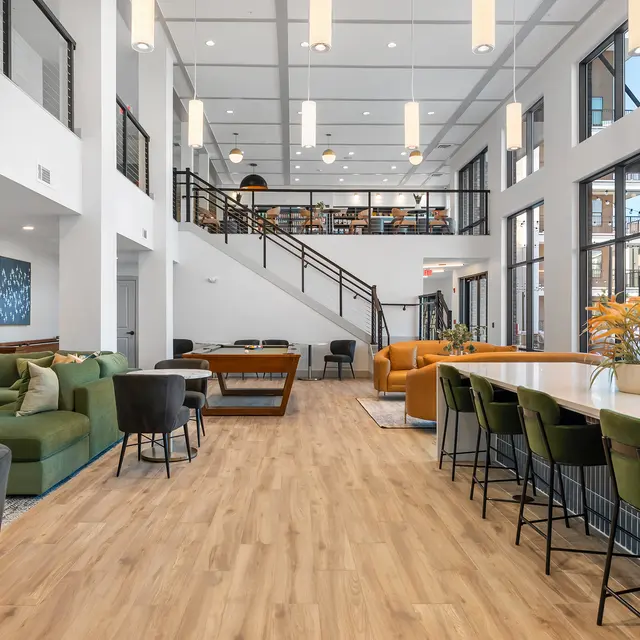 A spacious modern common area with comfortable seating, large windows, and a bright interior design. Features include green and orange furniture, a long table with chairs, and a staircase in the background.