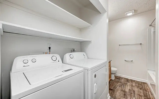 An interior view of a laundry room featuring a washing machine and dryer, with shelves above them and a bathroom visible in the background.