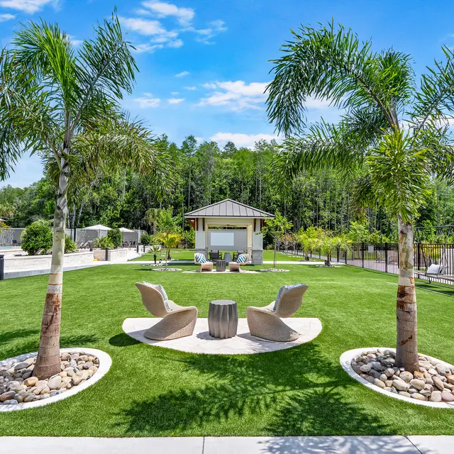 Outdoor Lounge Area A bright outdoor area featuring lush green grass, palm trees, and modern lounge chairs surrounding a central table. In the background, there's a small pavilion and landscaped surroundings with a wooden fence.