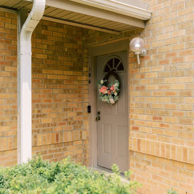 A residential front door with a decorative wreath, surrounded by a green bush and brick wall.