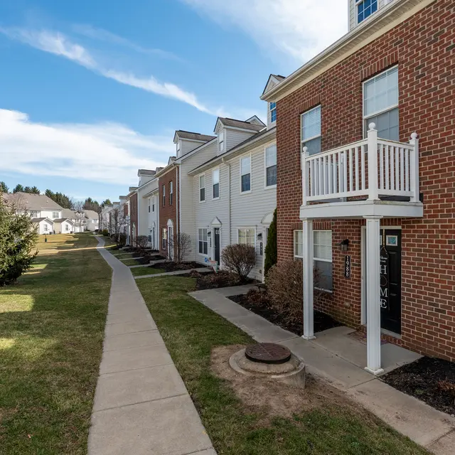 A residential neighborhood with a paved walkway along side several houses. The foreground features a brick house with a small balcony, while the background shows more houses of various colors. The sky is clear with some clouds, and there is greenery on either side of the path.