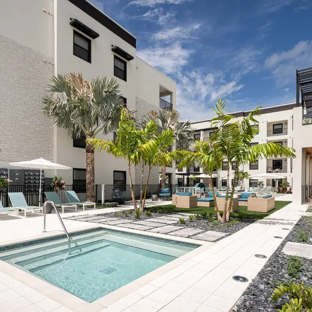 A modern apartment complex featuring a pool area with lounge chairs and tropical landscaping.