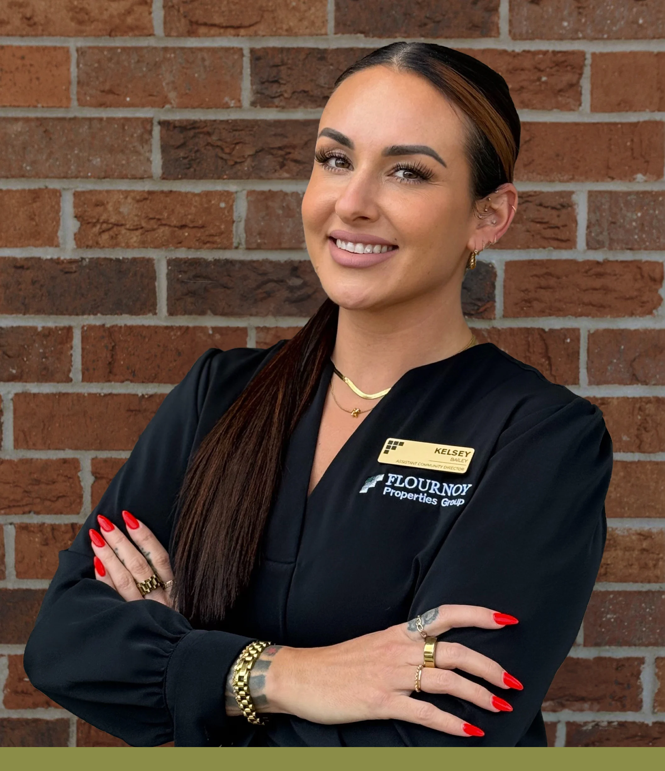Professional Real Estate Agent Portrait A smiling woman with long brunette hair and striking red nails stands confidently in front of a brick wall. She is wearing a black blouse with a name tag and is crossing her arms, giving a friendly impression.