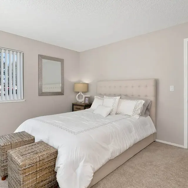 A cozy bedroom with a light-colored theme, featuring a bed with white bedding, two woven stools at the foot, and a nightstand with a lamp. A large window with blinds allows natural light, and there's a mirror on the wall.