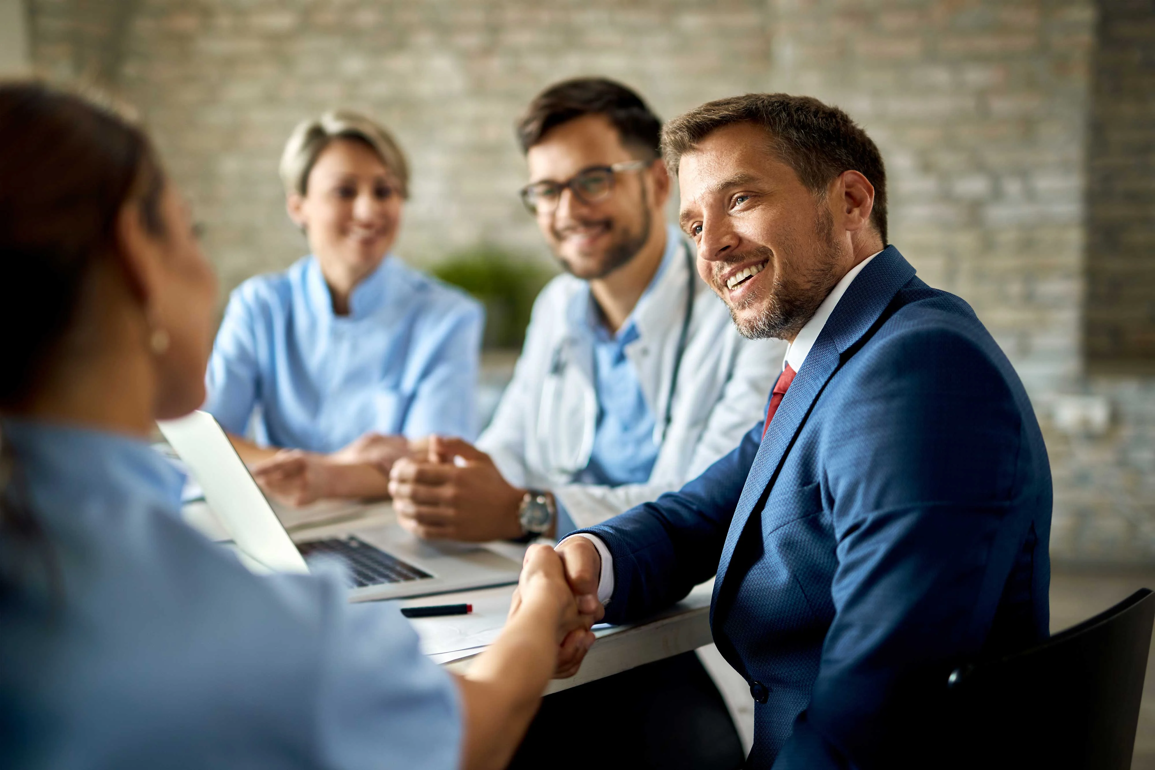 A business meeting with four professionals sitting at a table, smiling and engaging in discussion. One man in a suit is shaking hands with a woman, while two others are seated in the background looking pleased.
