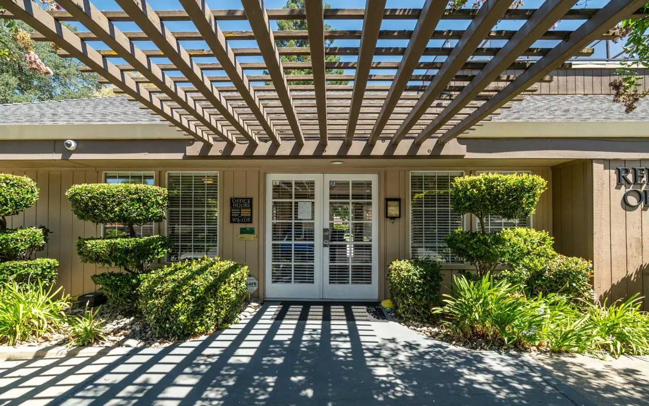 A building entrance with a wooden trellis above, flanked by neatly trimmed bushes and flowering plants.