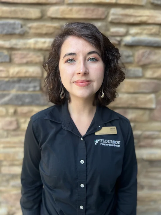 A young professional woman with curly hair is standing in front of a stone wall, wearing a black button-up shirt with a name tag and looking directly at the camera.