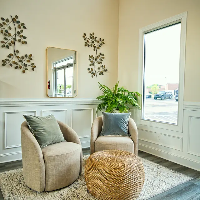 Cozy seating area with two rounded chairs, a knitted pouf, and decorative wall art in a well-lit room with large windows.