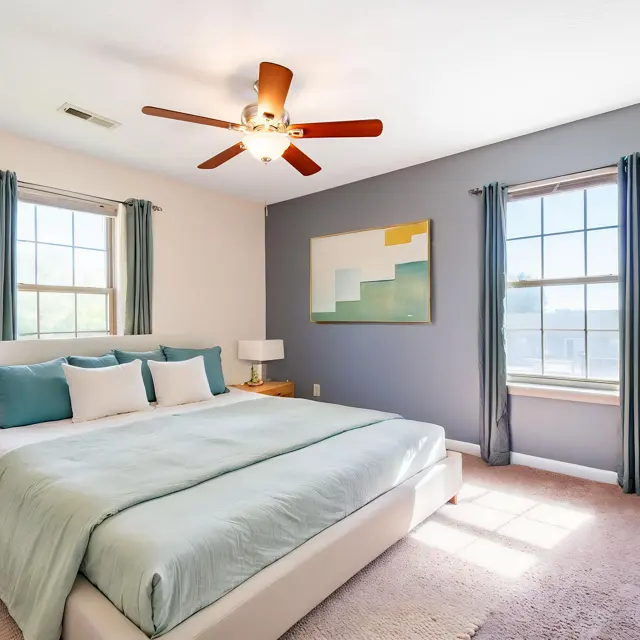 A modern bedroom featuring a large bed with a light blue comforter and several decorative pillows. The walls are painted in soft tones, with one wall having a dark gray color. There are two windows dressed with blue curtains, a ceiling fan, and a piece of abstract art on the wall. A potted plant and wooden furniture add warmth to the space.