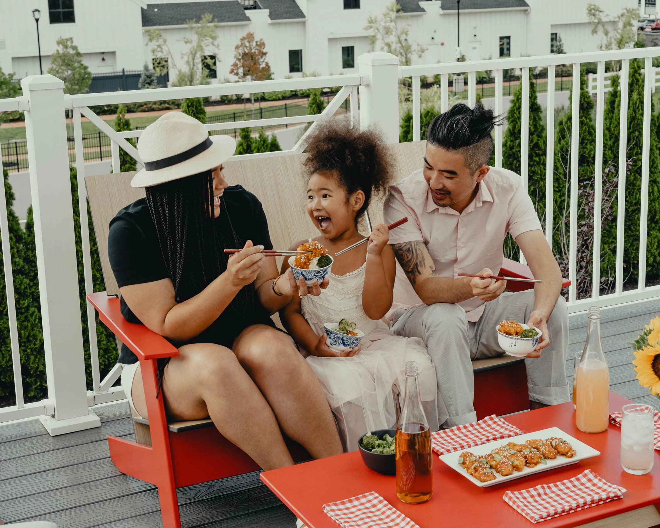 A family of three sits on a red outdoor sofa, enjoying food together. The child in the middle is laughing and holding a bowl. The adults are sharing food with the child, and there are drinks on the table.