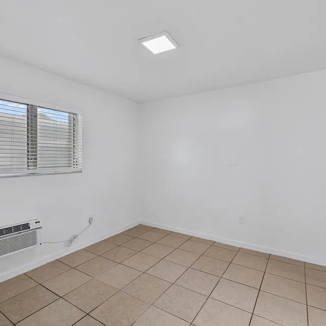 A bright, empty room with white walls and a tiled floor. There is a window with blinds on the left side and an air conditioning unit beneath it. The room has a simple ceiling light and an open doorway on the right leading to another area.