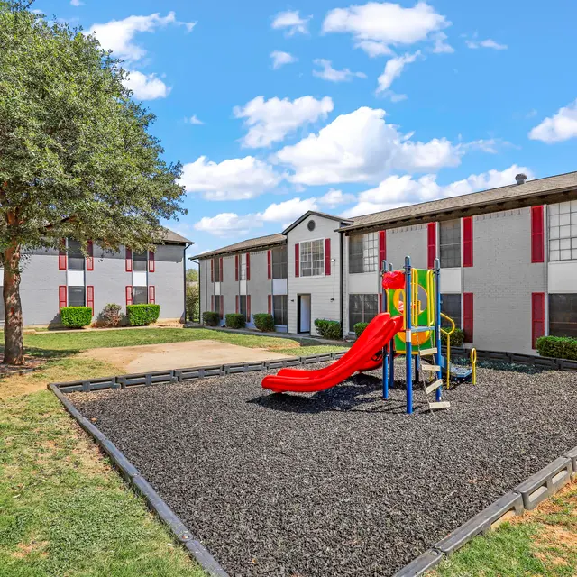 A playground featuring a colorful slide and climbing frame, located in the yard of an apartment complex with two-story buildings in the background, surrounded by grass and trees under a blue sky with fluffy clouds.