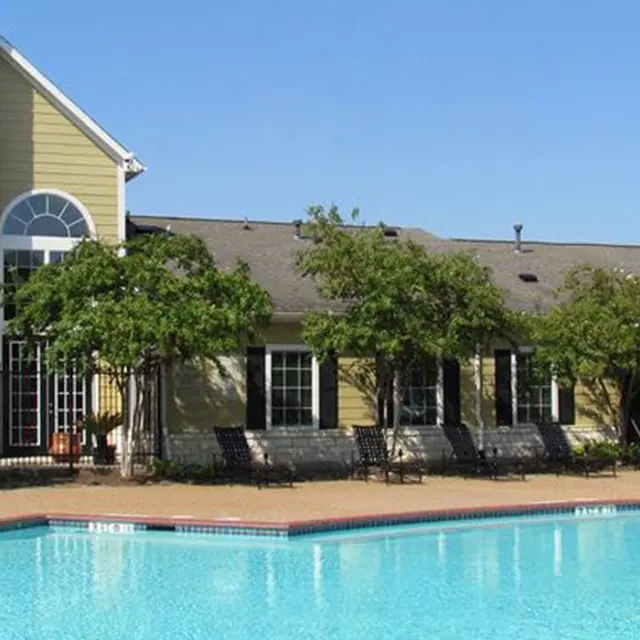 A clear blue swimming pool in front of a residential building with a stone chimney and large windows. Surrounding greenery and lounge chairs are visible.