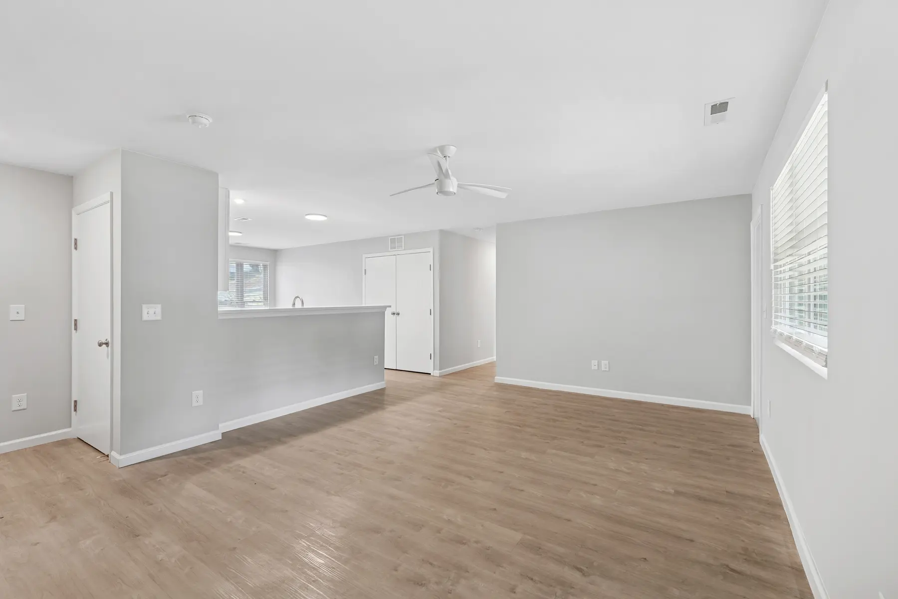 A spacious and bright living room featuring light-colored walls and wooden flooring, with a ceiling fan and a kitchen counter visible in the background.