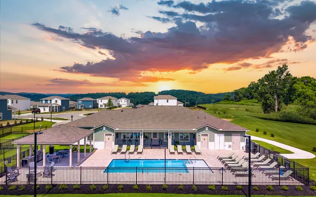 Aerial view of a residential pool area with a clear blue pool, surrounded by lounge chairs and a fenced lawn. In the background, houses are visible under a colorful sunset with dramatic clouds.