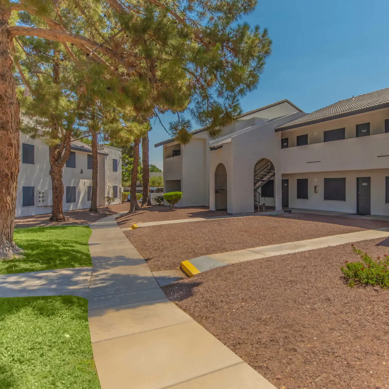 An exterior view of a quiet apartment complex featuring beige buildings and a landscaped courtyard with a walkway, trees, and gravel patches.