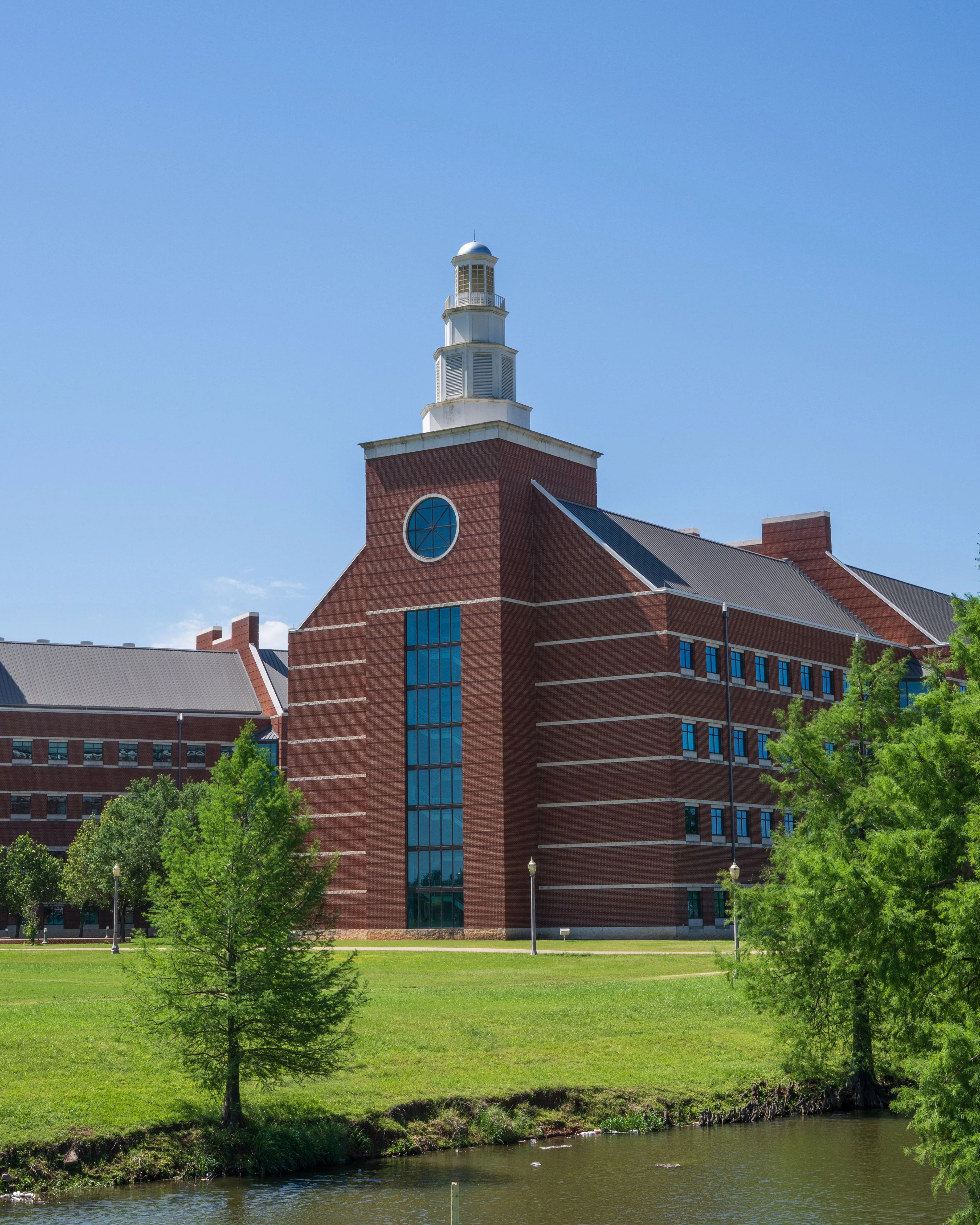 A large brick building with a dome at the top, surrounded by green grass and trees, beside a calm body of water under a clear blue sky.