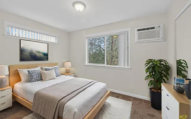 A cozy bedroom featuring a bed with decorative pillows, a window letting in natural light, a bedside table with lamps, a plant, and a piece of framed art on the wall.