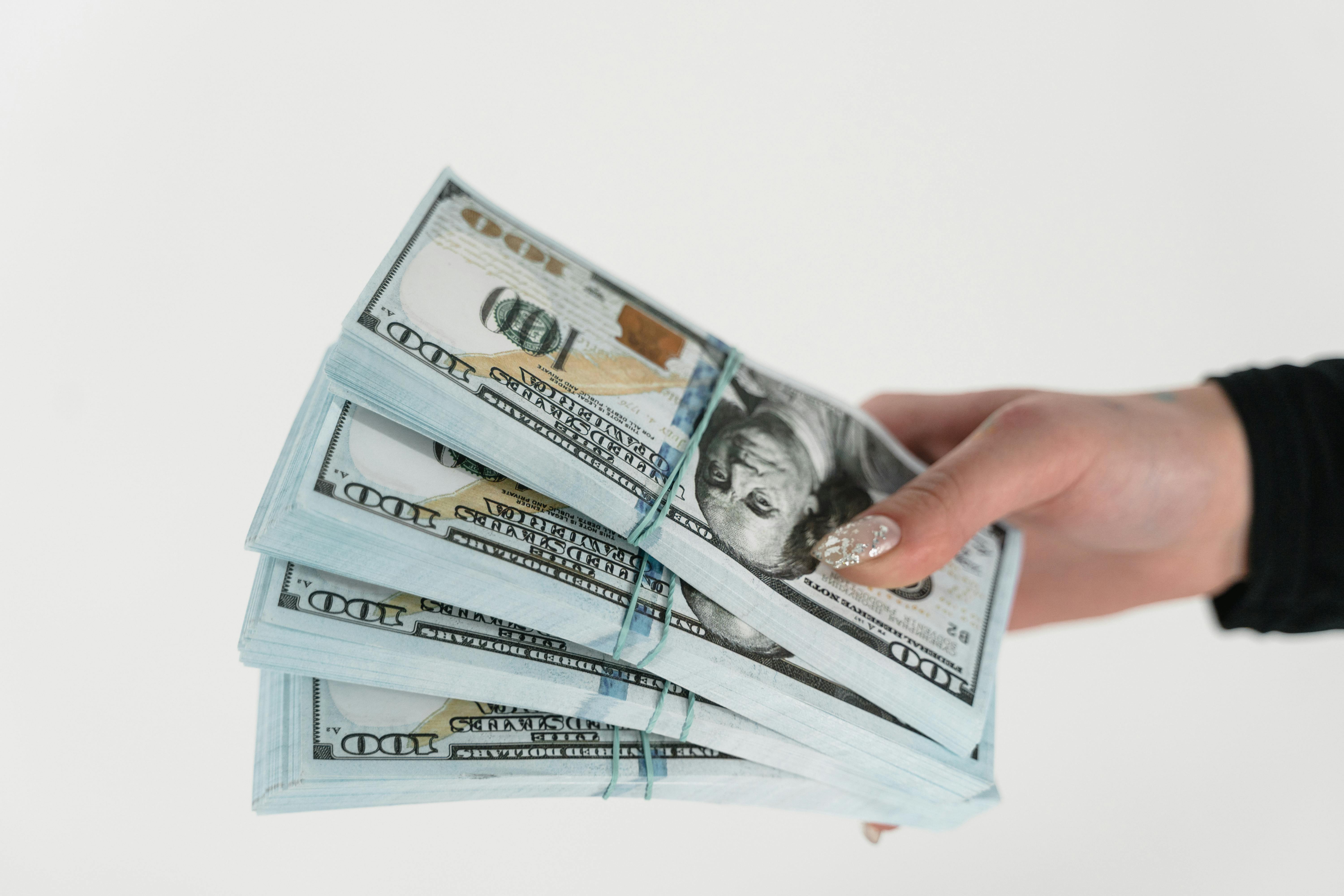 A hand holding several stacks of hundred-dollar bills secured with rubber bands.