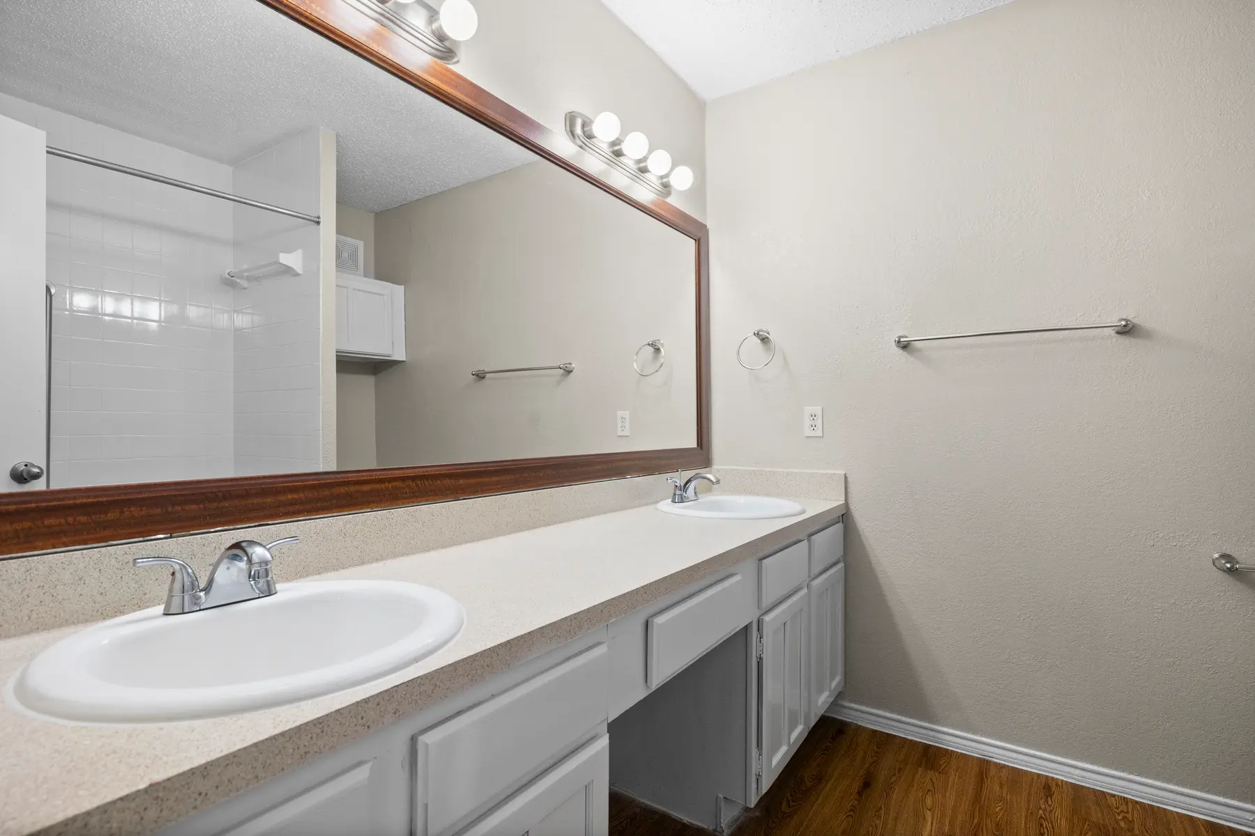 A modern bathroom featuring dual sinks with faucets on a light-colored countertop, a large mirror above, wooden flooring, and a separate shower area in the background.