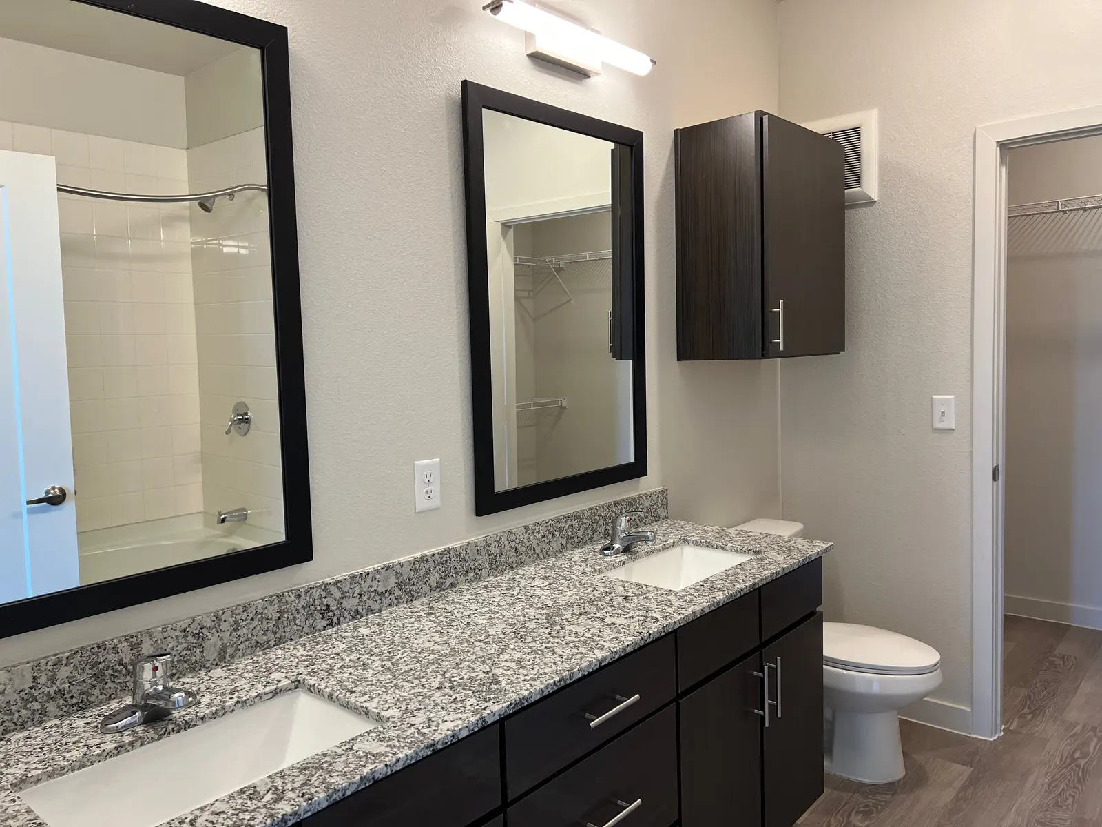 A modern bathroom featuring gray granite countertops, two sinks, large mirrors, dark cabinetry, and a white toilet in a clean space.