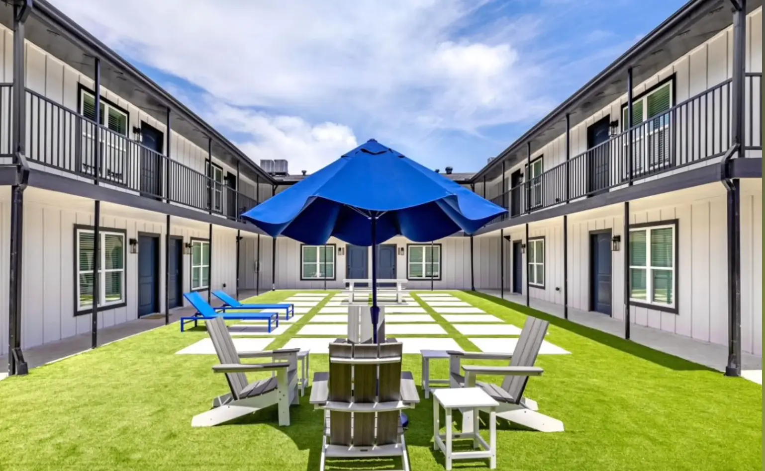 A scenic courtyard of a modern hotel featuring green grass, lounge chairs, and an umbrella. Surrounding the courtyard are two-story buildings with balconies.