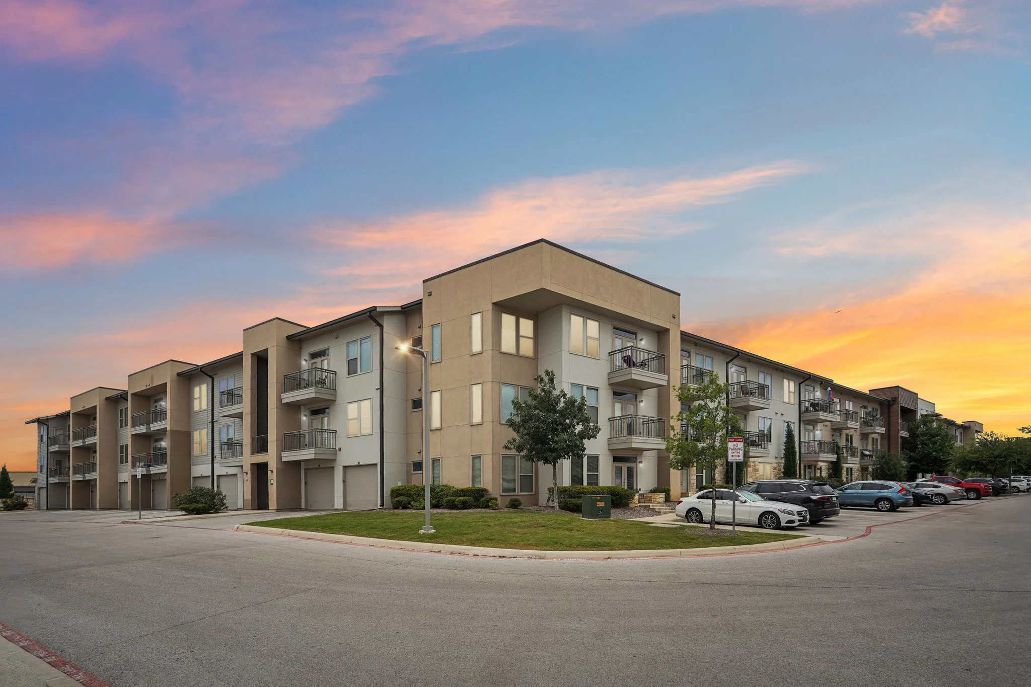 A modern apartment complex at dusk, featuring balconies and a well-kept lawn under a vibrant sunset sky.