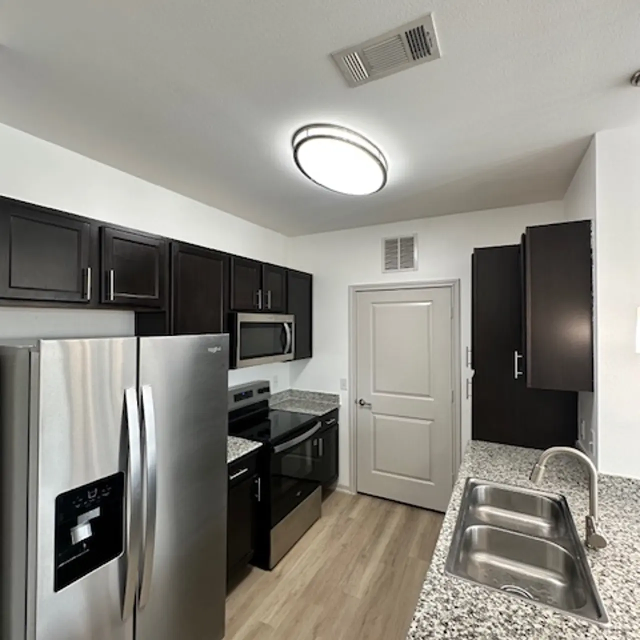 A modern kitchen with dark cabinets, stainless steel appliances, and a countertop made of speckled granite. The kitchen features a stove, microwave, and refrigerator, with a door leading to another room.
