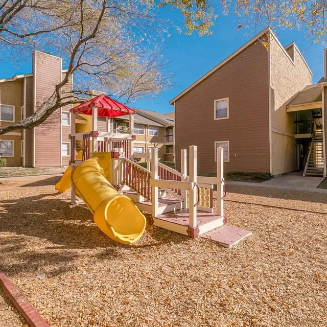 A children's playground featuring a yellow slide and play structure, surrounded by gravel and trees, with residential buildings in the background.