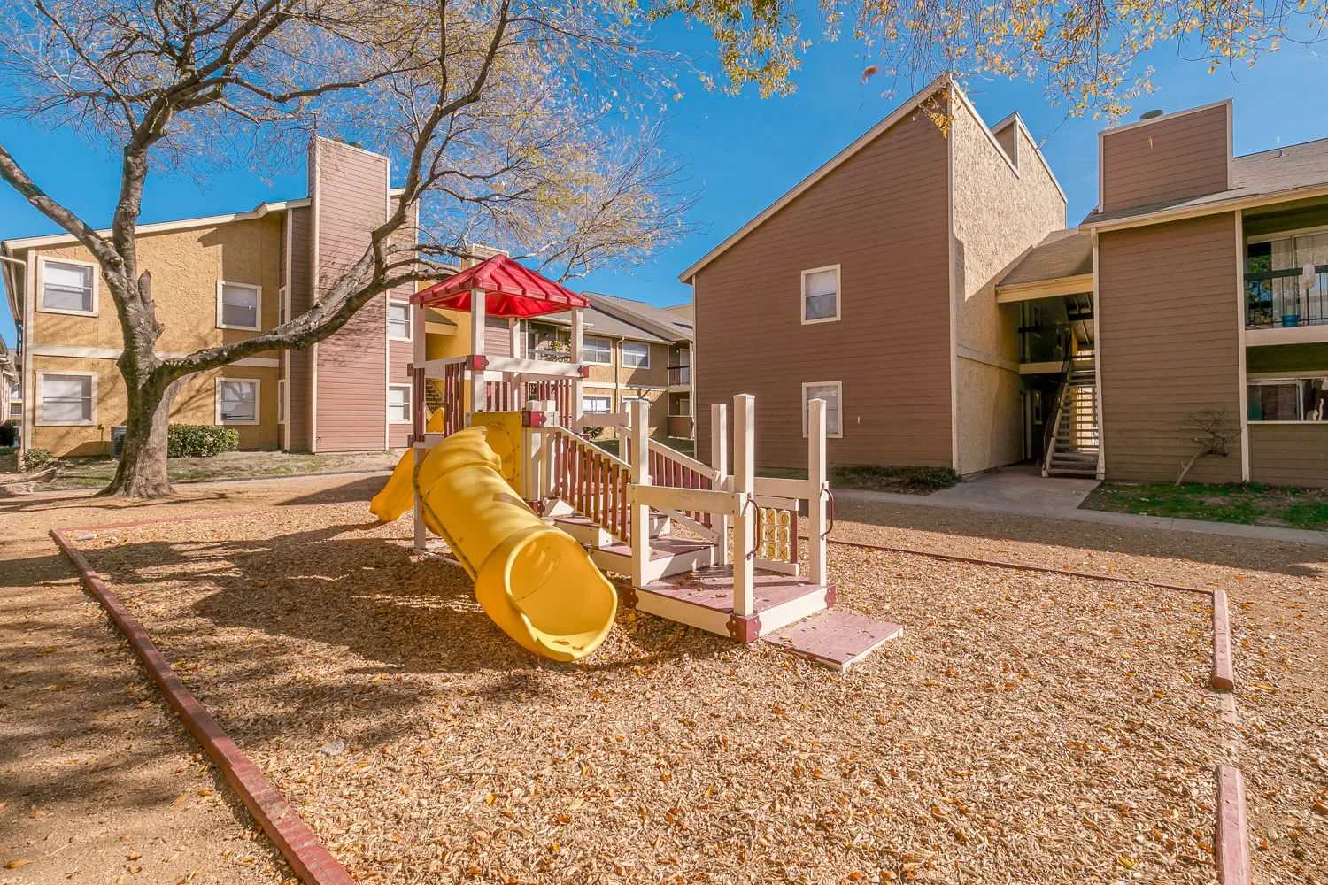 Children's Playground in Residential Area A children's playground featuring a yellow slide and play structure, surrounded by gravel and trees, with residential buildings in the background.