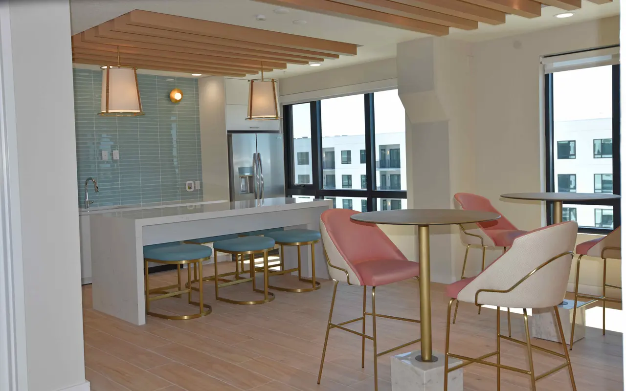 A modern kitchen area featuring light-colored wooden flooring, a white island with seating, and stylish pink and blue chairs. Large windows allow natural light to flood the space, highlighting the contemporary design elements.
